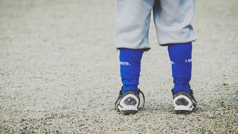 Image: Close-up of a child's legs and feet, clad in baseball gear, pointed away from us