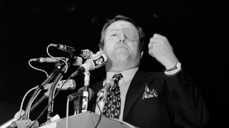 A younger eye-patched Le Pen with fist raised giving a speech