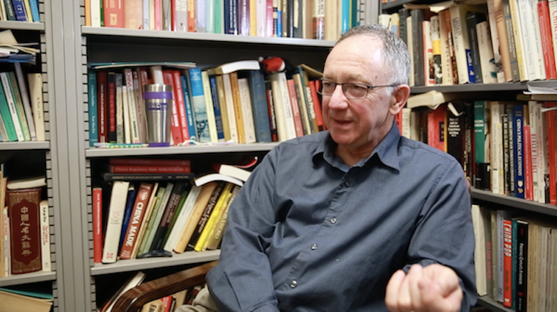 Tom Gold sitting in a chair, talking to someone off camera, with stuffed books shelves behind him